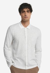 Polo shirt bianco a maniche lunghe con superficie texture, collarino strutturato e chiusura con bottoni frontali. Il tessuto appare leggero e morbido.