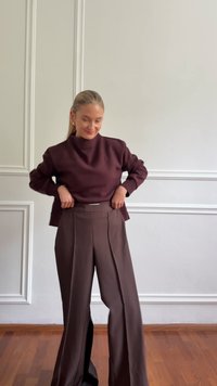 Vrouw met blond haar draagt een donkerpaarse top met lange mouwen en bijpassende hoge taille wijde broek, terwijl ze haar broek op haar taille ajusteert binnen.