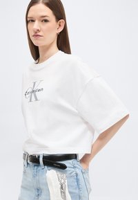Νεαρή γυναίκα με λευκό κοντομάνικο T-shirt Calvin Klein, ανοιχτόχρωμο τζιν, μαύρη ζώνη και ένα υφασμάτινο μοτίβο που κρέμεται από την τσέπη.