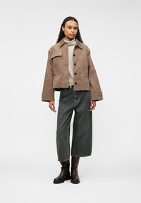 Chaqueta de pana marrón extragrande con bolsillo de solapa y cierre de botones, combinada con un suéter de punto gris y jeans oscuros de pierna ancha, finalizada con botas negras hasta el tobillo.