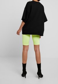 Maglietta nera oversize con maniche corte, abbinata a shorts da ciclismo verde neon brillante e stivaletti neri alla caviglia, vista da dietro.