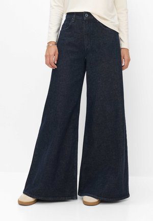 Person iført mørkeblå vidde jeans, en hvid langærmet top, beige sko og to tynde armbånd på det ene håndled.