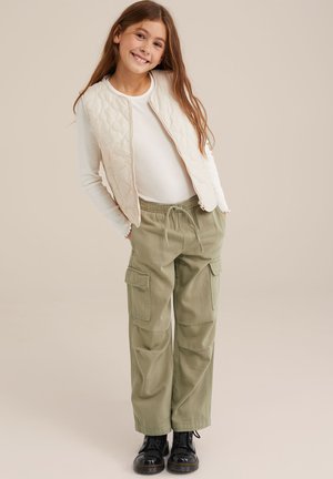 Chaleco de crema acolchado sobre una camiseta de manga larga blanca, combinado con pantalones cargo verdes oliva con bolsillos laterales y botas negras hasta el tobillo.