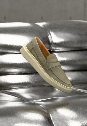 Light green suede slip-on shoe with a minimal design, cream rubber sole, and textured detailing on the side, set against a metallic background.