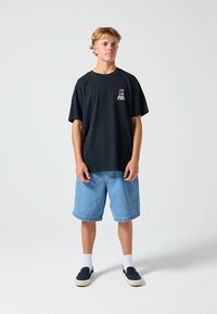 T-shirt preta de algodão com um pequeno logótipo floral, combinado com calções de ganga azul claro. O modelo usa sapatos slip-on azul-marinho e meias brancas.