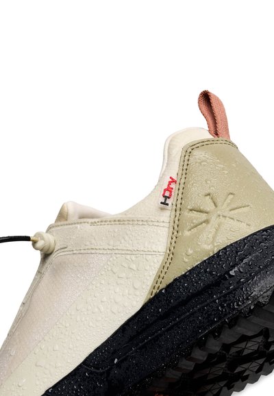 Gros plan sur une basket beige et noire avec semelle texturée, gouttes d'eau, détail d'onglet rouge et logo soleil brodé sur le talon.