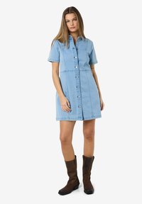 Vestido de ganga azul claro com mangas curtas, frente com botões e dois bolsos laterais. Usado com botas altas castanhas para contraste.