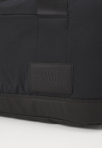 Black duffel bag featuring a textured, durable fabric with a flat rubberized logo patch. Includes a sturdy, webbed carrying strap.