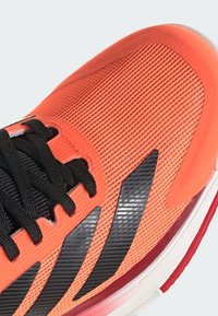 Sportovní obuv s texturovaným oranžovým síťovým svrškem, černými tkaničkami a reflexními černými detaily. Disponuje polstrovanou podešví v červené a bílé barvě.