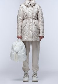 Prošívaný béžový svrchní oděv s vysokým límcem, doplněný bílým prošívaným batohem a šedými tepláky, zvýrazněný béžovými šněrovacími botami.