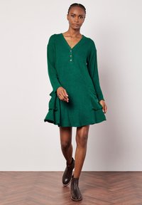 Abito in maglia verde a coste con scollo a V, maniche lunghe, patta con tre bottoni, orlo arricciato e tasche laterali, abbinato a stivali alla caviglia marroni scuri.