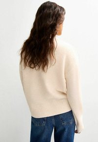 Jasno-beżowy sweter z prążkowanym wykończeniem, w luźnym kroju i długich rękawach, w połączeniu z niebieskimi dżinsami. Miękka faktura, minimalistyczny design.