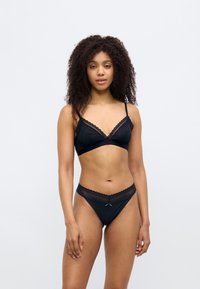 Σετ μαύρου εσωρούχου με bralette διακοσμημένο με δαντέλα και ρυθμιζόμενες τιράντες, συνοδευόμενο από τόνγκα με κυματιστά άκρα και μικρή διακοσμητική φιογκάκι.