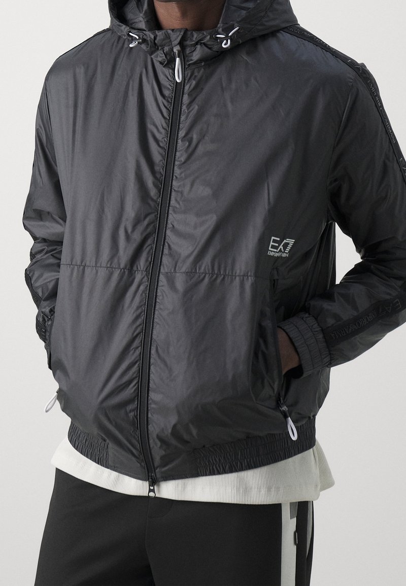 Chaqueta con capucha negra con cremallera y puños y dobladillo elásticos, logo de EA7 Emporio Armani en el pecho y las mangas, llevada sobre una camisa blanca.