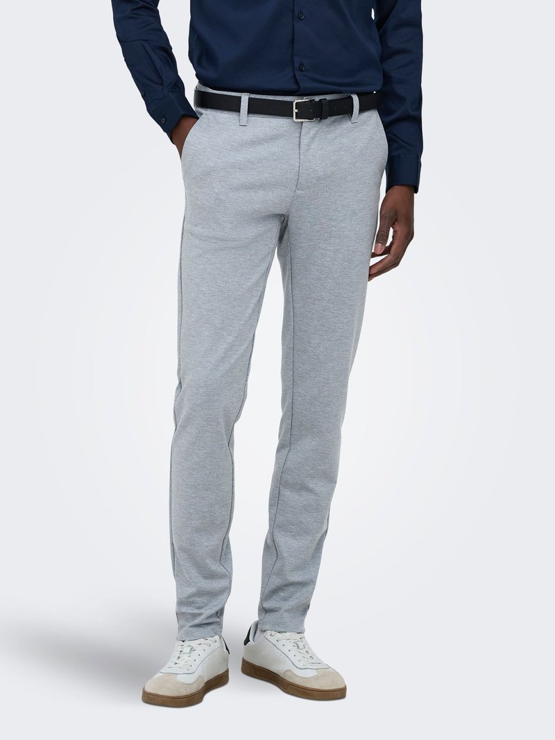 Homme portant un pantalon slim gris clair, ceinture noire, chemise bleu marine boutonnée et baskets blanches, debout avec une main dans la poche.