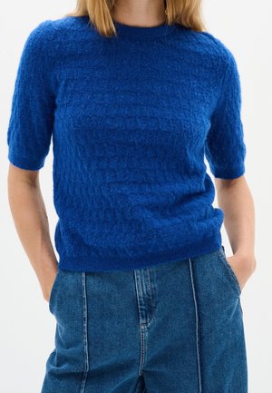 Kvinde iført en struktureret kongeblå kortærmet trøje og højtaljede blå jeans med hænderne i lommerne mod en ensfarvet baggrund.