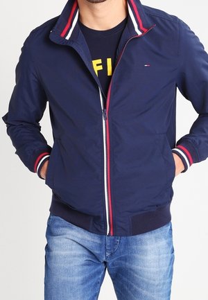 Homme portant une veste zippée bleu marine avec un col et des poignets à rayures rouges, blanches et bleues, associée à un jean bleu clair.