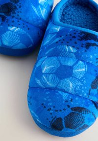 Blå fleeceslippers med ett mönster av fotbollar, med en plyschinteriör och solid blåa detaljer, rund form och mjuk textur.