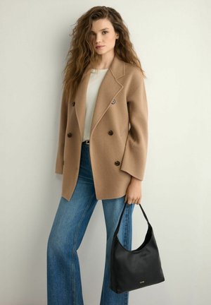 Kobieta z długimi kręconymi włosami nosi dwurzędowy płaszcz w kolorze wielbłądzim, biały top, niebieskie dżinsy i trzyma czarną torebkę na jednolitym jasnym tle.