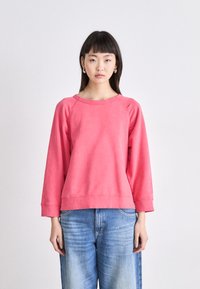 Sudadera rosa claro de corte relajado, con mangas raglán, escote redondeado y puños de canalé, combinada con jeans de mezclilla.