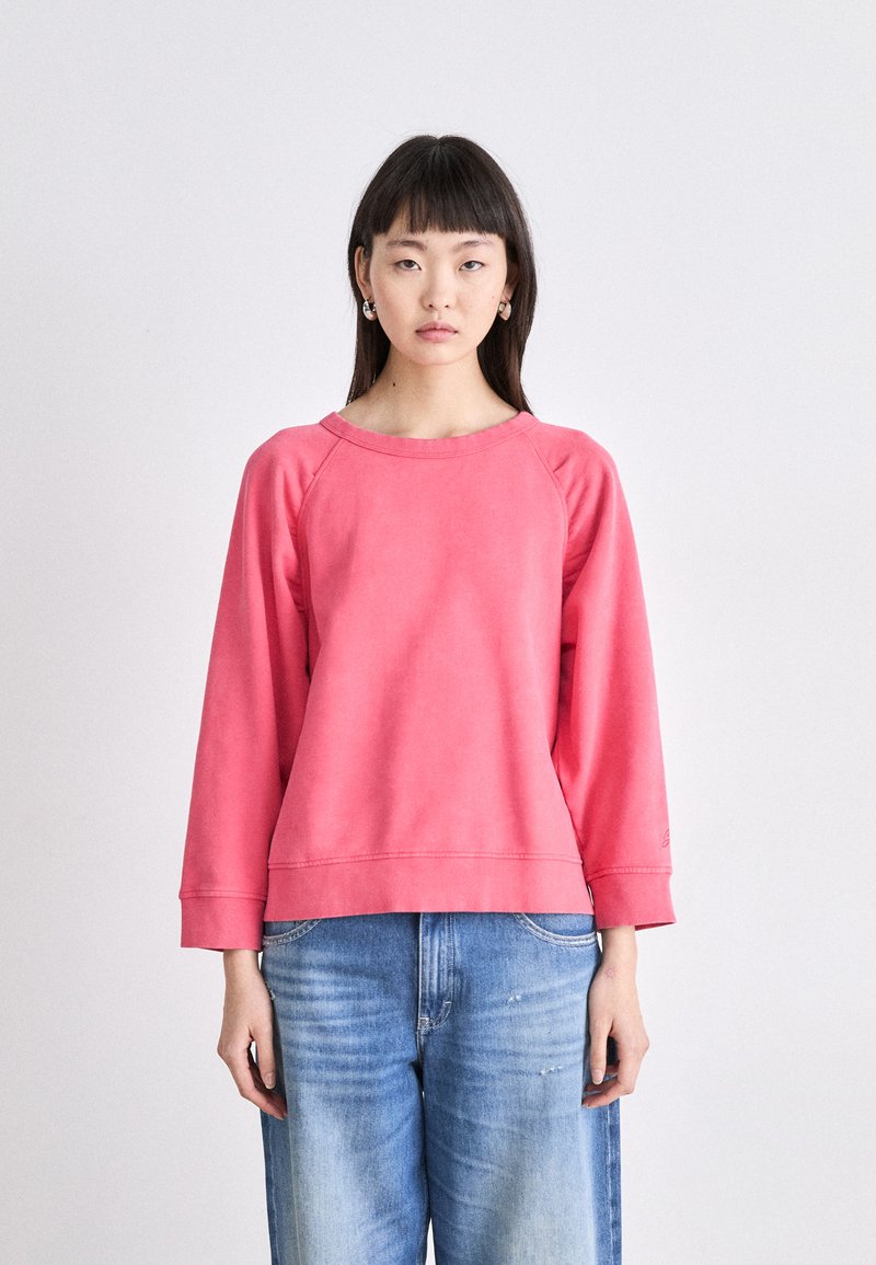 Sudadera rosa claro de corte relajado, con mangas raglán, escote redondeado y puños de canalé, combinada con jeans de mezclilla.