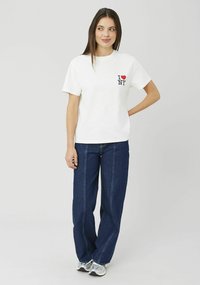 T-shirt en coton blanc avec un cœur rouge et du texte noir "I NY" sur le côté gauche de la poitrine, assorti à un jean large bleu foncé et des baskets argentées.