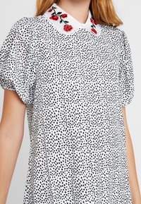 White blouse with black polka dots, puff sleeves, and a white collar featuring red floral embroidery. Smooth fabric texture.