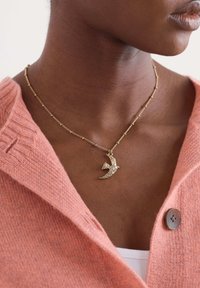 Arany nyaklánc, madár formájú medállal, texturált dizájnnal; rózsaszín kötött pulóver fölött viselve, gomb részletekkel.