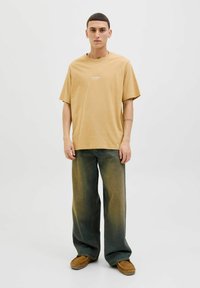 T-shirt en coton beige à manches courtes, présentant un petit logo blanc. Associé à un jean vert clair large et décoloré et des chaussures à enfiler marron.