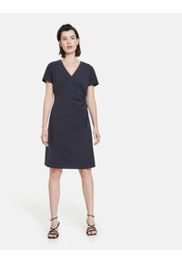 Marineblaues Kleid mit V-Ausschnitt, kurzen Ärmeln und einem leicht drapierten Design. Glatte Textur, knielang, kombiniert mit schwarzen Riemchensandalen.