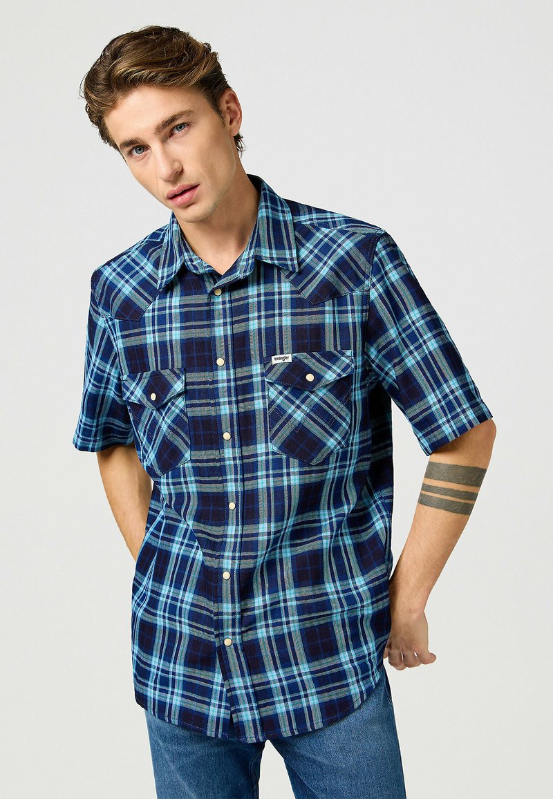 Camicia a maniche corte a quadri con motivi a check blu e blu navy, due tasche sul petto e chiusura con bottoni. Materiale in cotone dalla texture liscia.