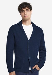 Marinblå stickad kofta med sjalkrage, utrustad med två framfickor och knappstängning. Strukturerad tyg ger djup.