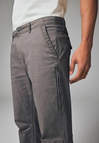 Graue Cargo-Hosen aus weichem Stoff, mit Seitentaschen und einer Reißverschlusstasche. Gerades Bein mit sichtbaren Nahtdetails.
