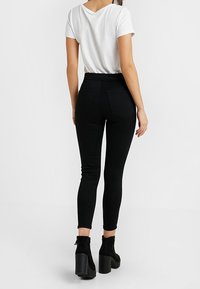 Kvinna som bär en vit kortärmad topp, höga svarta skinny jeans och svarta klackboots, stående med ryggen vänd mot kameran.