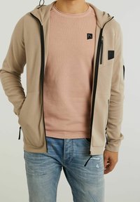 Felpa con zip beige in tessuto testurizzato, con accenti neri e una toppa con logo; indossata sopra un maglione rosa chiaro a girocollo. Jeans blu visibili.