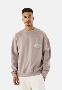 Sudadera marrón con cuello redondo, mangas largas y un estampado gráfico en el lado izquierdo del pecho que presenta texto y un emblema. Textura suave.