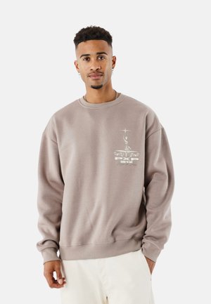 Sudadera marrón con cuello redondo, mangas largas y un estampado gráfico en el lado izquierdo del pecho que presenta texto y un emblema. Textura suave.