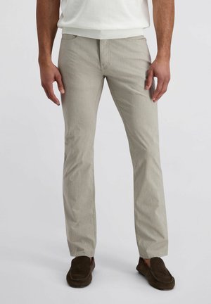 Man in lichtbeige rechte broek en bruine instapschoenen, staand tegen een effen witte achtergrond.