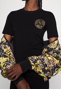 Svart kortärmad t-shirt med en gyllene cirkulär logotyp. Har en mönstrad pufferjacka i svart, guld och vitt som tillbehör.