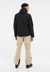 Chaqueta aislante negra con capucha, combinada con pantalones de nieve beige. Detalles notables incluyen un logo en la chaqueta y cremalleras decorativas en los pantalones.