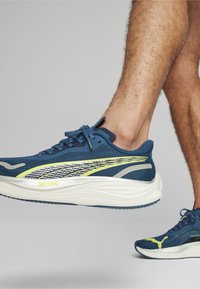 Scarpe sportive blu con tomaia in rete, dettagli riflettenti argento, accenti gialli e suola bianca spessa. Design con lacci per una vestibilità sicura.