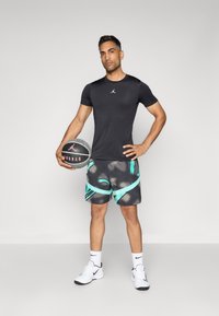 Melnā sporta T-kreklā ar mazu logo, pelēkās biksēs ar abstraktiem tirkīza rakstiem, turēdams basketbola bumbu. Baltās sporta apavās.