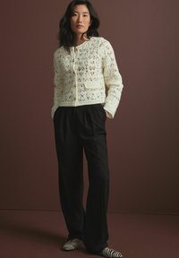 Gehaakte crèmekleurige cardigan met een open kantpatroon, voorzien van gouden knopen en een klein zakje, gecombineerd met zwarte losse broeken en gestreepte schoenen.