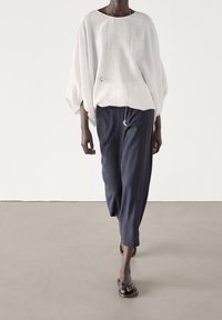 Top bianco in lino oversize con maniche a palloncino, abbinato a pantaloni blu scuro a gamba larga e sandali neri. Design minimalista.