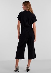 Tuta nera con maniche corte e vita elasticizzata, caratterizzata da gambe ampie e cropped. Indossata con scarpe con tacco a righe bianche e nere.