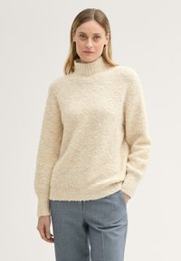 Sweter w kolorze kremowym z wysokim kołnierzem, wykonany z puszystej, strukturalnej tkaniny. Posiada długie rękawy i luźny krój, zestawiony z szarymi spodniami.