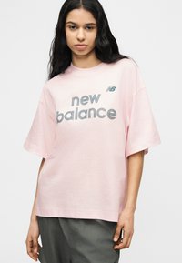 Νέα γυναίκα με μακριά σκούρα μαλλιά φοράει ένα φαρδύ ανοιχτό ροζ t-shirt New Balance και σκούρο παντελόνι, στέκεται μπροστά σε ένα απλό φωτεινό φόντο.