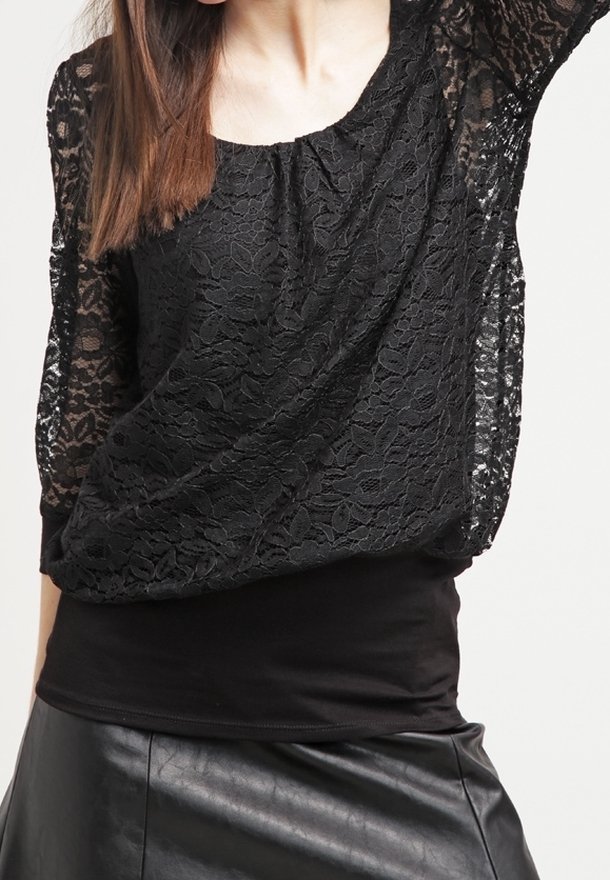 Femme portant un haut en dentelle noire avec manches transparentes et une jupe en cuir noire, cadrée du cou aux hanches sur fond blanc.