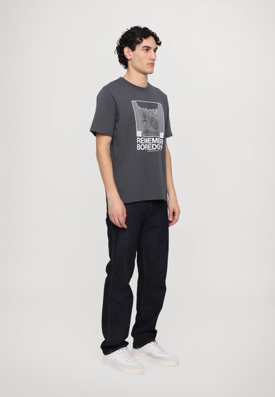 Gray cotton t-shirt with a graphic print, paired with dark denim jeans. Footwear is white sneakers with a patterned sole.