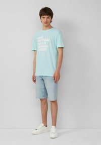 Lichtblauwe T-shirt met witte grafische tekst, gecombineerd met lichtkleurige denim korte broek en witte sneakers. De stof is zacht en casual.